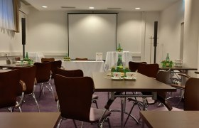 Seminarraum mit Tischen, Stühlen und Getränken, Projektionsleinwand im Hintergrund.